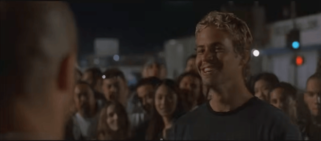 A crowd gathering at night, with a young man smiling and interacting with someone out of view. The scene captures excitement and engagement among the onlookers.
