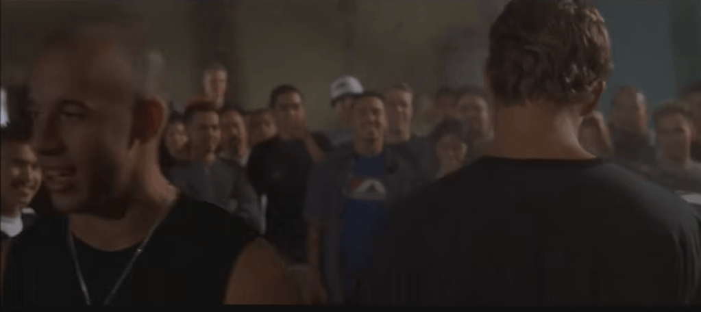 A scene featuring two male characters, one facing forward with a bald head and the other with a muscular build and medium-length hair, surrounded by an audience in a dimly lit space.