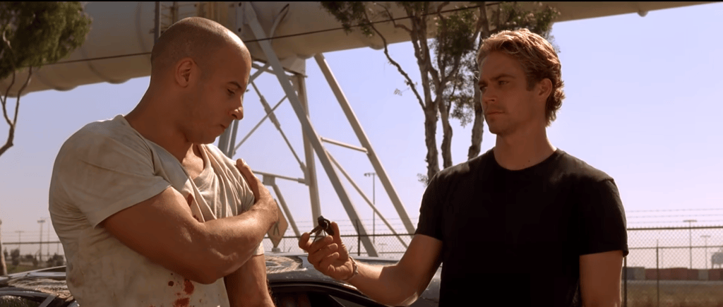 Two men standing near a car, one with a shaved head wearing a dirty shirt and the other with blonde hair in a black t-shirt, exchanging a small object.